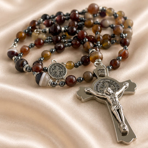 A beaded rosary with brown agate beads, a silver St Benedict medallion, and a silver crucifix, on a draped champagne satin fabric.