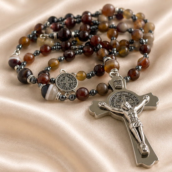 A beaded rosary with brown agate beads, a silver St Benedict medallion, and a silver crucifix, on a draped champagne satin fabric.