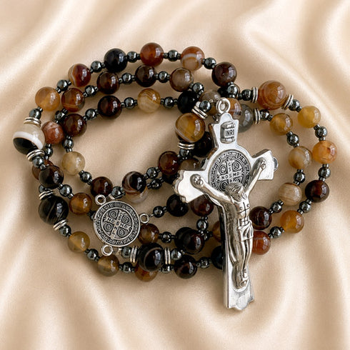 A rosary with brown and dark beads, a silver-tone St Benedict crucifix and a medallion, on a beige satin fabric.