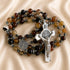 A rosary with brown and dark beads, a silver-tone St Benedict crucifix and a medallion, on a beige satin fabric.