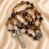 A rosary with brown beads, a silver-tone St Benedict crucifix and a medal, on a beige satin fabric.