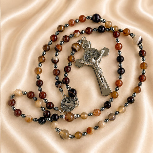 A rosary with brown, black, and beige agate beads, a silver crucifix and a medal, on a draped beige satin fabric background.