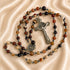 A rosary with brown, black, and beige agate beads, a silver crucifix and a medal, on a draped beige satin fabric background.