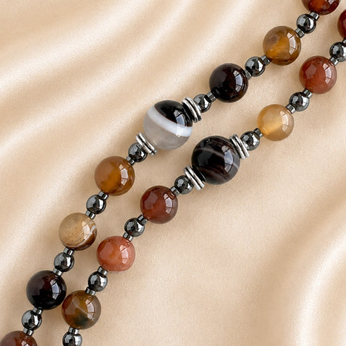 A close-up of a brown, amber, orange, black, and white stones beaded necklace on a soft beige satin fabric.