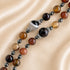 A close-up of a brown, amber, orange, black, and white stones beaded necklace on a soft beige satin fabric.
