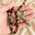 A close-up of a rosary with brown and beige stone beads, a silver crucifix, and a medal, held in a hand against a beige background.