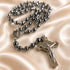 Hematite rosary with St Benedict medallion, and a silver-toned crucifix, on a cream satin fabric background.