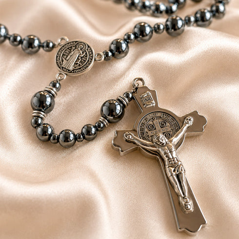 A close-up of a rosary with dark metallic beads and silver-toned medallions and crucifix, arranged on draped satin fabric.