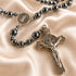 A close-up of a rosary with dark metallic beads and silver-toned medallions and crucifix, arranged on draped satin fabric.