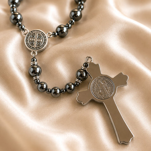 A rosary with dark hematite beads and a silver-tone cross pendant with a St Benedict medallion, on a champagne satin fabric.