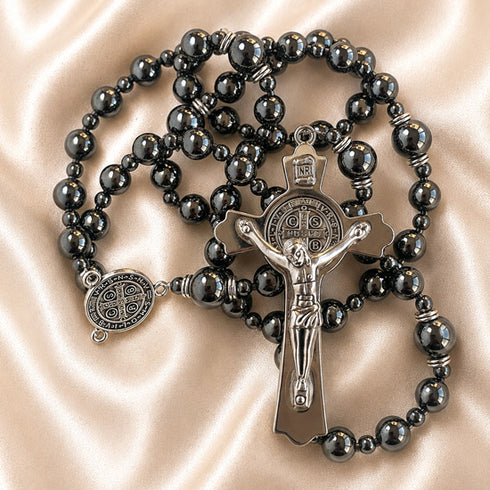 A black beaded rosary with a silver-tone crucifix and St Benedict medallion, on a beige satin fabric.