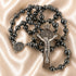 A black beaded rosary with a silver-tone crucifix and St Benedict medallion, on a beige satin fabric.