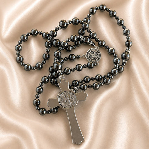 A hematite rosary with a silver-toned cross pendant and a round medallion, arranged on a satin fabric background.