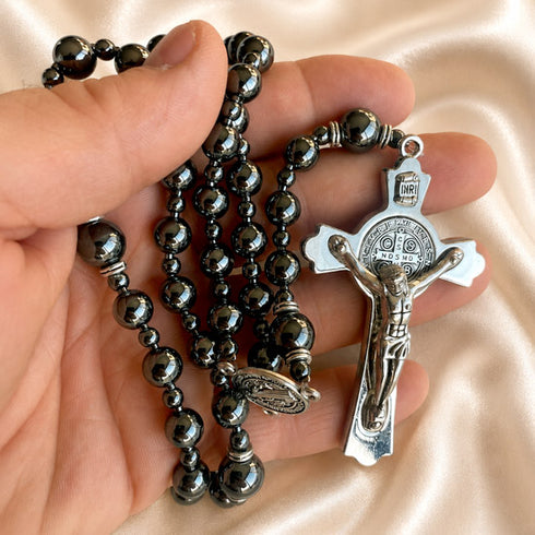 A hand holding a black hematite rosary with a silver crucifix against a satin cream fabric background.