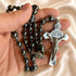 A hand holding a black hematite rosary with a silver crucifix against a satin cream fabric background.