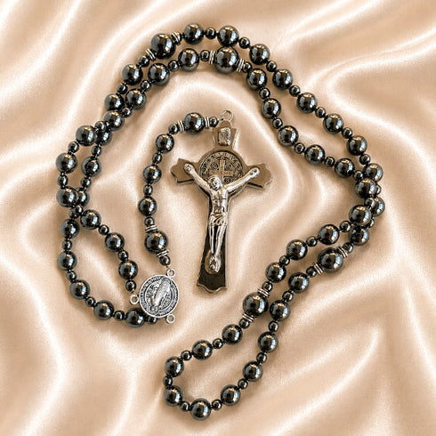 A rosary with hematite beads, a silver-toned crucifix, and a small round St Benedict medal, on a satin fabric background.