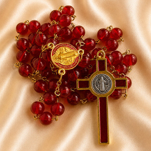 A red glass bead rosary with a gold-tone and silver-tone St Benedict cross, on a beige satin background.