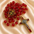 A red glass bead rosary with a gold-tone crucifix and centerpiece medal, arranged on a satin fabric background.