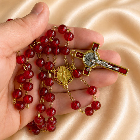 A close-up of a hand holding a red and gold rosary, a gold crucifix, and a gold medallion, on a beige satin background.