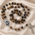 A rosary with brown tiger's eye beads, a round silver medal, and a silver cross, on a beige satin fabric background.
