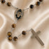 A rosary with brown tiger's eye beads, a St. Charbel medal, and a silver-tone cross, displayed on a satin background.