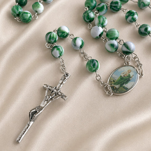 A close-up of a marbled green and white rosary laid on a beige satin fabric background.