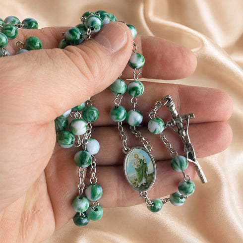 A close-up of a hand holding a green and white rosary with a silver crucifix and an oval centerpiece, on a soft beige satin.