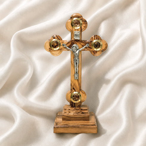 A frontal image of a hand-crafted small standing olive wood crucifix with halo and intricate details, featuring olive leaves, dried flowers, holy soil and incense on white background.