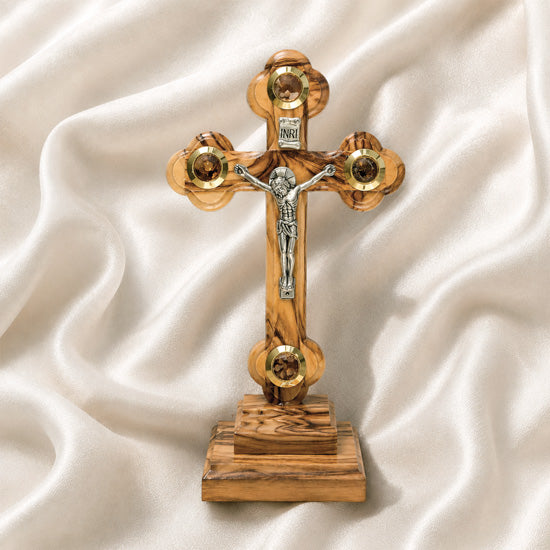 A frontal image of a hand-crafted small standing olive wood crucifix with halo and intricate details, featuring olive leaves, dried flowers, holy soil and incense on white background.