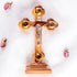 A frontal image of a hand-crafted small standing olive wood crucifix with halo and intricate details, featuring olive leaves, dried flowers, holy soil and incense on white background.