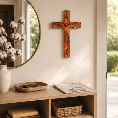 Wooden cross on a wall above a wooden console table with a vase, mirror, and newspaper.