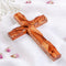 A handcrafted olive wood cross with a natural brown finish, placed on a white fabric with scattered pink petals around it.