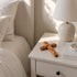 Wooden cross on a nightstand next to a bed with white bedding and a lamp.