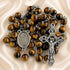 A rosary with polished brown tiger's eye beads, a silver-tone crucifix, and a central oval medallion with the text 'Ruega por Nosotros'.