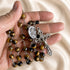 A close-up of a hand holding a rosary with brown and black tiger's eye beads, a medal of St Charbel, and a crucifix.