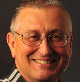Smiling older man with glasses on dark grey background.