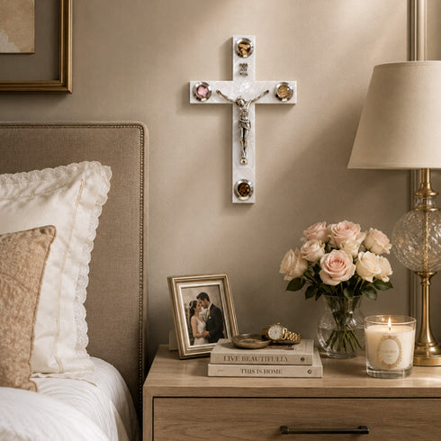 White latin cross on a wall above a nightstand with books, flowers, and a lamp.