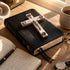 Black leather-bound Bible with a decorative cross on a wooden table, surrounded by a notebook, pen, and cup.