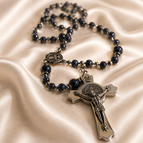 A dark blue lapis lazuli rosary with silver-toned metal beads, a St Benedict medallion, and a crucifix, on a beige satin background.