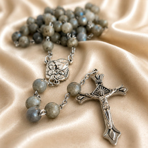 A close-up of a rosary with gray labradorite beads and silver-tone metal components, on draped champagne satin fabric.