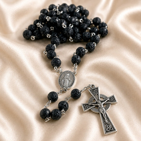 A black lava stone rosary with silver metal links, a round medallion, and a detailed crucifix pendant, on a satin background.