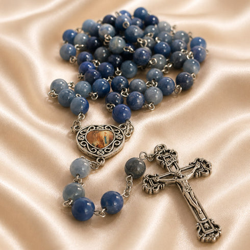 A rosary with blue beads, a silver-tone crucifix, and a heart-shaped centerpiece with, on a satin fabric background.